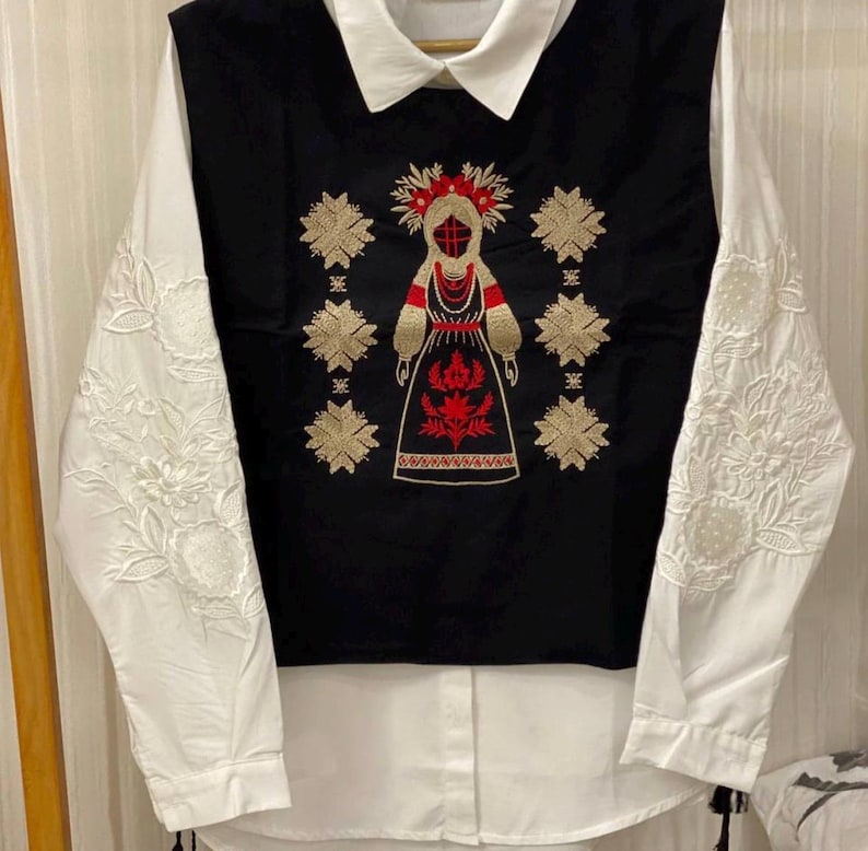 May include: A long-sleeved white shirt featuring a black vest overlay. The black section displays intricate gold and red embroidery of a central figure surrounded by geometric star patterns. The white sleeves are adorned with detailed white floral embroidery.