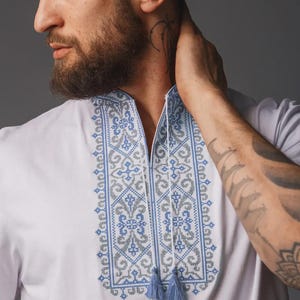 May include: A white shirt featuring a vertical panel of intricate blue and grey cross-stitch embroidery down the center. The neckline has a split opening with two thin white cords ending in small blue tassels hanging from the collar area.