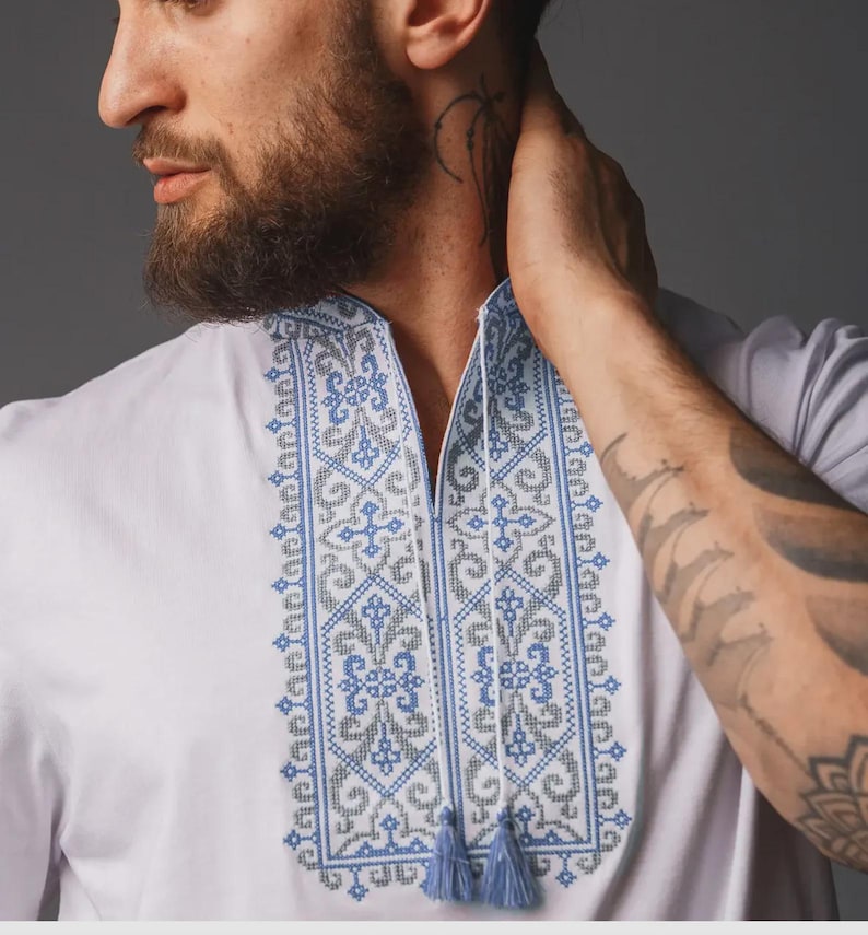 May include: A white shirt featuring a vertical panel of intricate blue and grey cross-stitch embroidery down the center. The neckline has a split opening with two thin white cords ending in small blue tassels hanging from the collar area.
