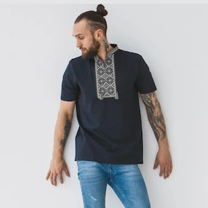 May include: A navy blue short-sleeved shirt with an embroidered geometric design on the collar. The shirt is paired with light blue denim jeans. The shirt's design features white and gray patterns.