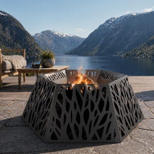 Op de afbeelding: Een zwarte, geometrische vuurkorf met een bladachtig uitgesneden ontwerp. De vuurkorf is gevuld met brandend hout en staat op een stenen terras. De achtergrond toont een meer, bergen en een houten terras met tuinmeubelen.