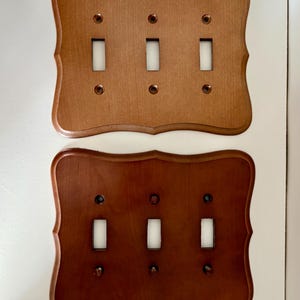 May include: Two rectangular wooden light switch plates with scalloped edges. Each plate features three vertical rectangular cutouts for switches and six small circular holes for mounting screws. One plate is light brown, the other is dark brown.