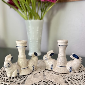 May include: Cream-colored ceramic bunny candle holders with blue accents. Each holder features a bunny figurine and a candlestick holder. The bunnies have blue ears and small blue flower details. The candle holders are on a lace doily.