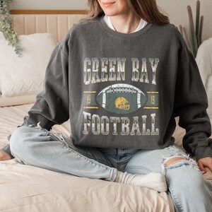 May include: Dark grey sweatshirt featuring the text "GREEN BAY FOOTBALL" in a distressed style. The design includes a football and helmet graphic. The sweatshirt is worn with light blue jeans.