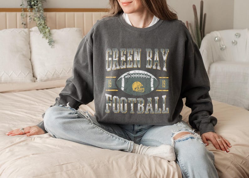 May include: Dark grey sweatshirt featuring the text "GREEN BAY FOOTBALL" in a distressed style. The design includes a football and helmet graphic. The sweatshirt is worn with light blue jeans.