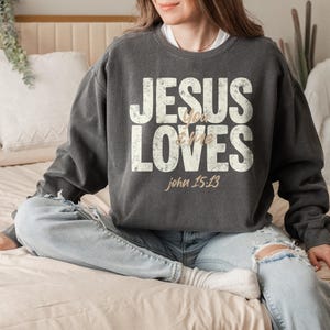 May include: Dark gray sweatshirt with the text "JESUS loves me" in distressed white lettering. The Bible verse John 15:13 is printed below in a light brown color. The sweatshirt is a casual style.