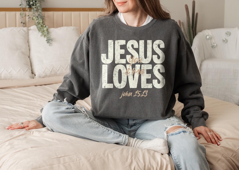 May include: Dark gray sweatshirt with the text "JESUS loves me" in distressed white lettering. The Bible verse John 15:13 is printed below in a light brown color. The sweatshirt is a casual style.