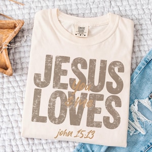 May include: Cream-colored t-shirt with the text "JESUS LOVES you" in distressed brown lettering. The text "john 15:13" is printed in a smaller, gold-colored font. The shirt is folded on a white surface.