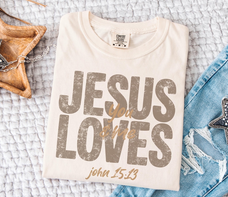 May include: Cream-colored t-shirt with the text "JESUS LOVES you" in distressed brown lettering. The text "john 15:13" is printed in a smaller, gold-colored font. The shirt is folded on a white surface.