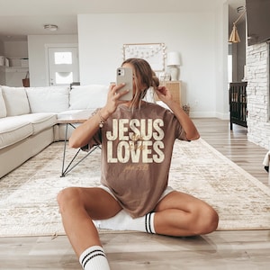 May include: A brown t-shirt with the text "JESUS LOVES" in a distressed, vintage style. The text "John 3:16" is below the main text. The shirt is worn with white shorts and socks with black stripes.