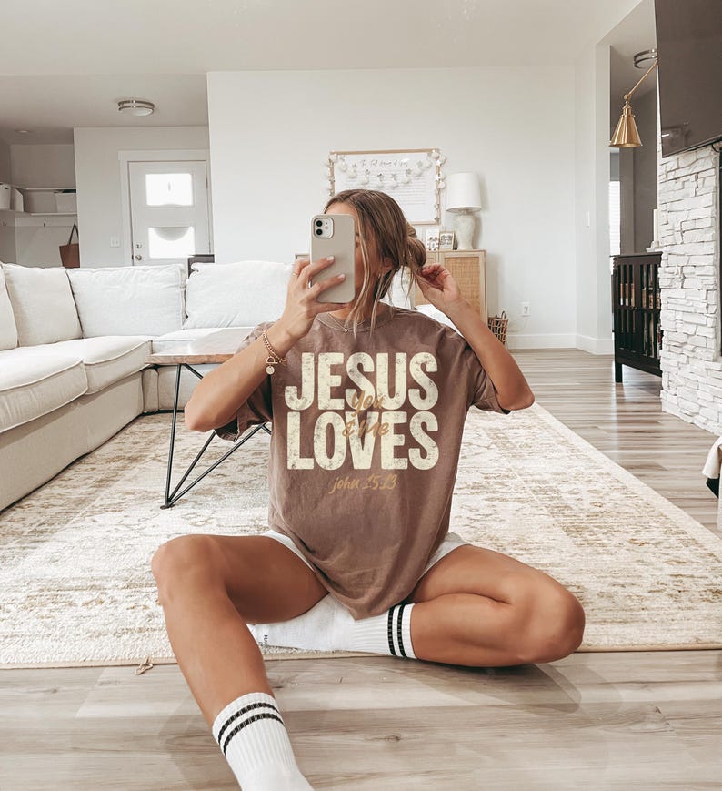 May include: A brown t-shirt with the text "JESUS LOVES" in a distressed, vintage style. The text "John 3:16" is below the main text. The shirt is worn with white shorts and socks with black stripes.