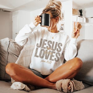 May include: White sweatshirt with the text "JESUS LOVES" in a distressed gold font, with "John 15:13" below. The person is holding a black mug. The person is wearing slippers and sitting on a gray couch.