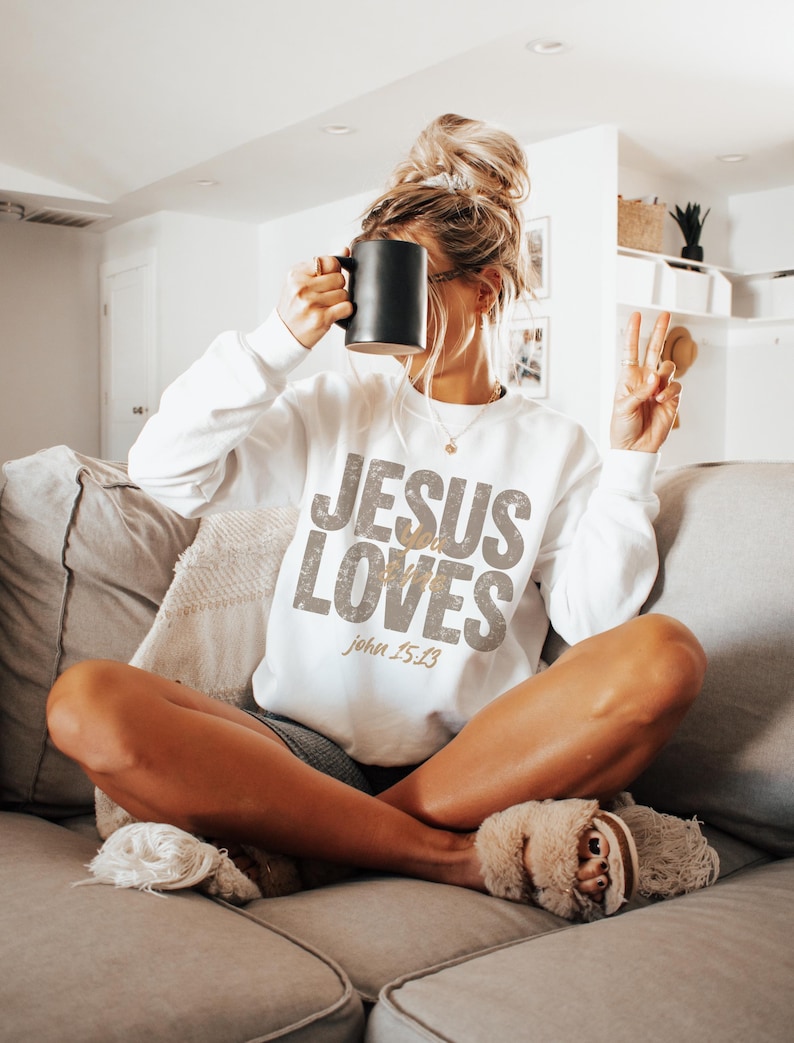 May include: White sweatshirt with the text "JESUS LOVES" in a distressed gold font, with "John 15:13" below. The person is holding a black mug. The person is wearing slippers and sitting on a gray couch.