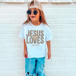May include: A young person wearing a white t-shirt with the text "JESUS LOVES" in a distressed gold font, with "John 15:13" below. The person is also wearing blue jeans, a baseball cap, and sunglasses.