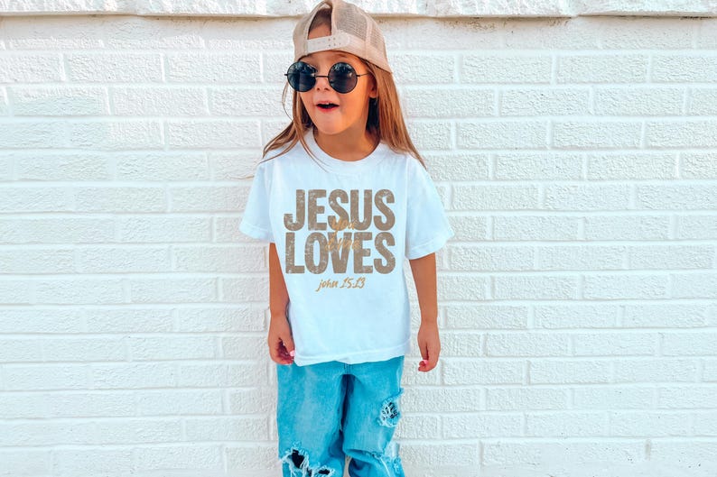 May include: A young person wearing a white t-shirt with the text "JESUS LOVES" in a distressed gold font, with "John 15:13" below. The person is also wearing blue jeans, a baseball cap, and sunglasses.