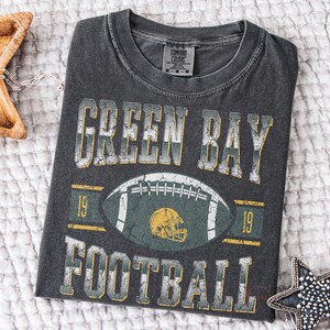 May include: Dark grey t-shirt featuring the text "GREEN BAY FOOTBALL" in distressed white and yellow lettering. A football graphic with a green and yellow helmet is centered on the shirt. The t-shirt is folded on a white textured surface.