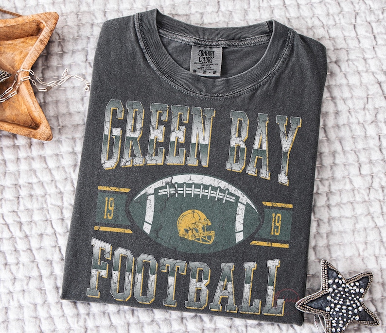 May include: Dark grey t-shirt featuring the text "GREEN BAY FOOTBALL" in distressed white and yellow lettering. A football graphic with a green and yellow helmet is centered on the shirt. The t-shirt is folded on a white textured surface.