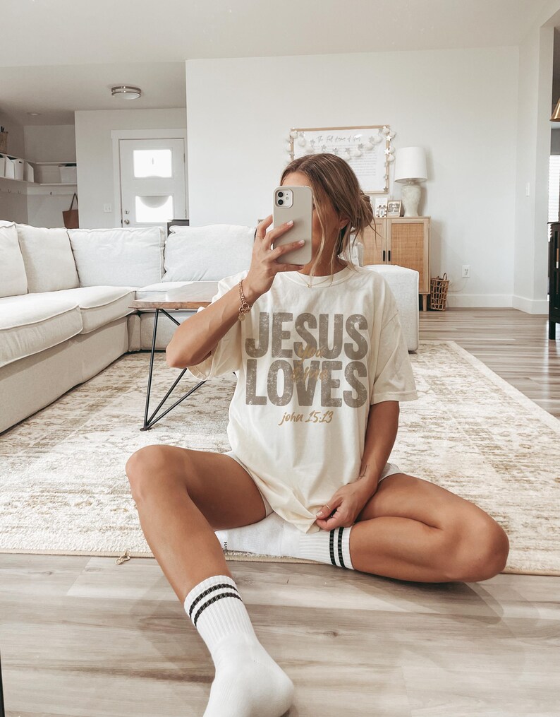 May include: Cream-colored t-shirt with the text "JESUS LOVES" in a distressed, textured font. The text "John 15:13" is below. The person is wearing white socks with black stripes. The shirt is worn with the person sitting on a rug.