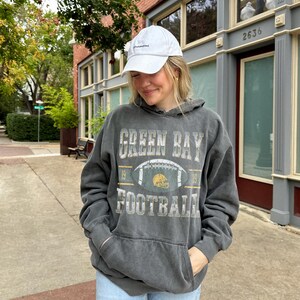 May include: A person wearing a white baseball cap and a dark grey Green Bay Football hoodie with a football graphic. The person is also wearing light blue jeans. The hoodie has a front pocket and the text "Green Bay Football" printed on the front.