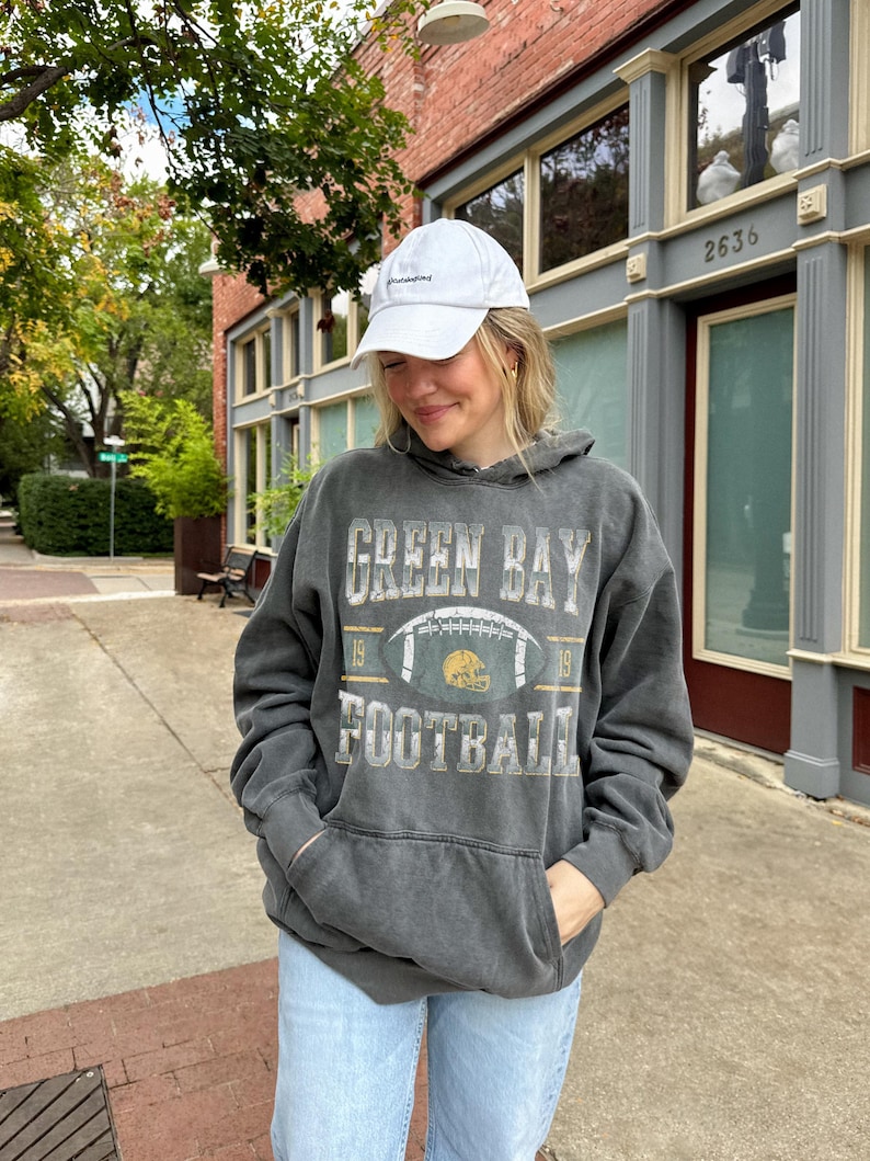 May include: A person wearing a white baseball cap and a dark grey Green Bay Football hoodie with a football graphic. The person is also wearing light blue jeans. The hoodie has a front pocket and the text "Green Bay Football" printed on the front.