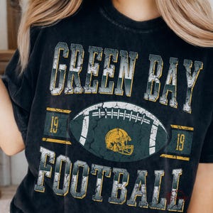 May include: Black t-shirt with a distressed graphic design. The design features the words "GREEN BAY FOOTBALL" above and below a football and a helmet. The numbers "19" are on either side of the football.