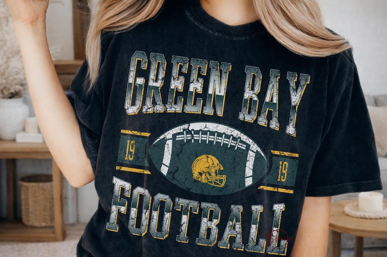May include: Black t-shirt with a distressed graphic design. The design features the words "GREEN BAY FOOTBALL" above and below a football and a helmet. The numbers "19" are on either side of the football.