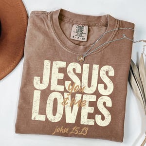 May include: A brown t-shirt with the text "JESUS loves you & me" in a distressed, off-white font. The text "john 15:13" is below. A silver necklace with a small pendant rests on the shirt. A brown hat and dried leaves are in the background.
