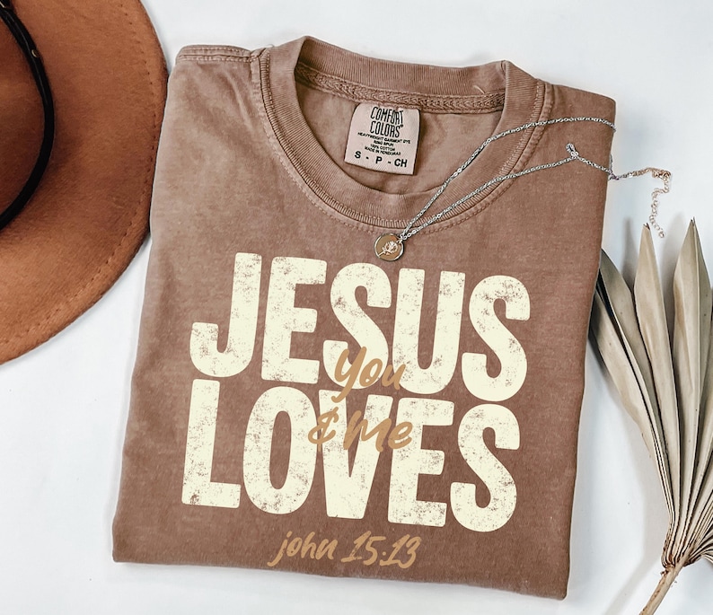 May include: A brown t-shirt with the text "JESUS loves you & me" in a distressed, off-white font. The text "john 15:13" is below. A silver necklace with a small pendant rests on the shirt. A brown hat and dried leaves are in the background.
