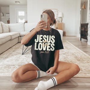 May include: Black t-shirt with the text "JESUS LOVES" in white, with "John 15:13" in gold. The shirt is worn with white shorts and white socks with black stripes. The person is holding a phone.