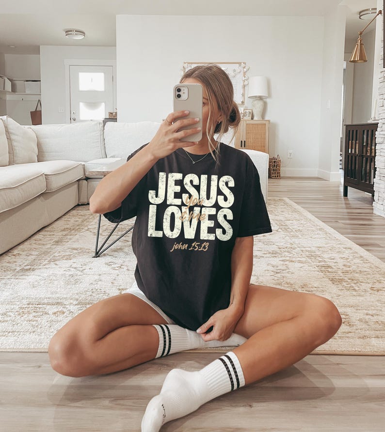 May include: Black t-shirt with the text "JESUS LOVES" in white, with "John 15:13" in gold. The shirt is worn with white shorts and white socks with black stripes. The person is holding a phone.