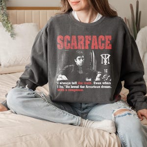 K&ouml;nnte beinhalten: Dunkelgraues Sweatshirt mit dem Wort "SCARFACE" in Rot und einem Schwarz-Wei&szlig;-Bild eines Mannes. Das Sweatshirt enth&auml;lt auch das Zitat: "Ich sage immer die Wahrheit. Auch wenn ich l&uuml;ge. Er liebte den amerikanischen Traum. Mit Rache."