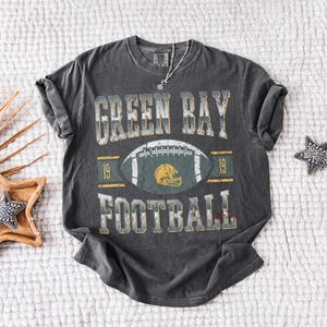 May include: Dark grey t-shirt featuring the text "GREEN BAY FOOTBALL" and a football graphic. The design includes a football helmet and the number "19". The shirt has a vintage, distressed appearance.