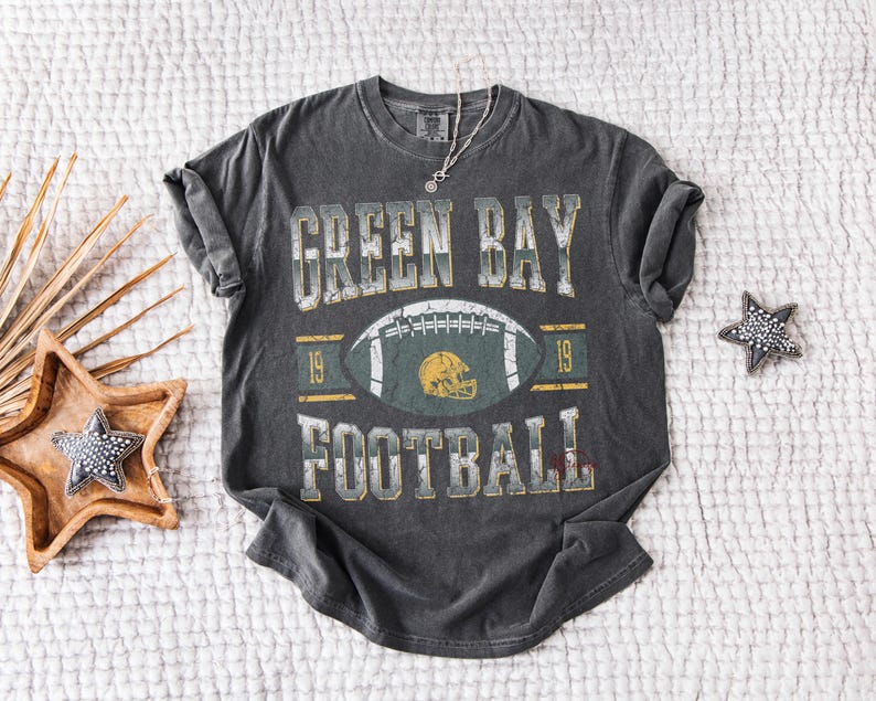 May include: Dark grey t-shirt featuring the text "GREEN BAY FOOTBALL" and a football graphic. The design includes a football helmet and the number "19". The shirt has a vintage, distressed appearance.