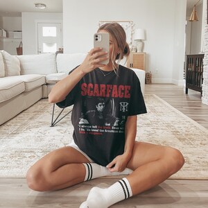 K&ouml;nnte beinhalten: Schwarzes T-Shirt mit dem Wort "SCARFACE" in roten Blockbuchstaben. Das Shirt zeigt ein Schwarz-Wei&szlig;-Bild eines Mannes und den Text "I always tell the truth. Even with I lie. He loved the American dream with a vengeance."