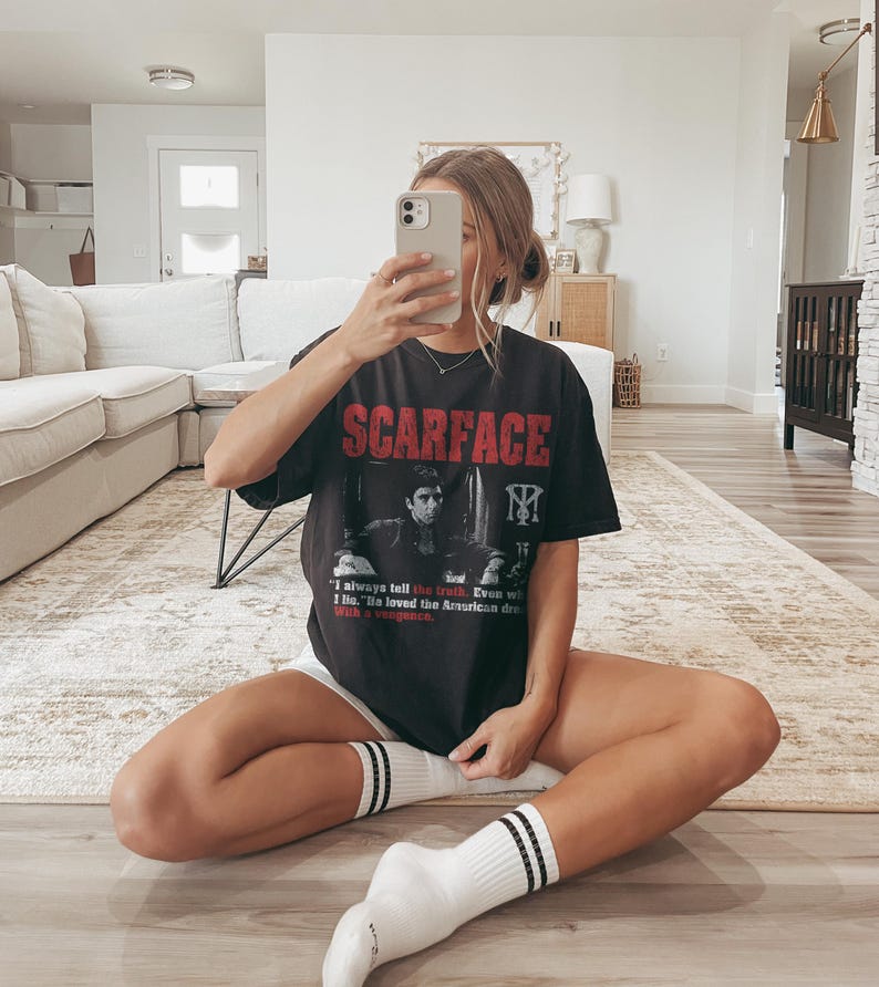 K&ouml;nnte beinhalten: Schwarzes T-Shirt mit dem Wort "SCARFACE" in roten Blockbuchstaben. Das Shirt zeigt ein Schwarz-Wei&szlig;-Bild eines Mannes und den Text "I always tell the truth. Even with I lie. He loved the American dream with a vengeance."