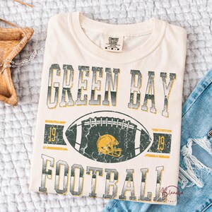 May include: Cream-coloured t-shirt featuring the text "GREEN BAY FOOTBALL" in a distressed, vintage style. A football graphic with a yellow helmet is centered below the text. The shirt has a classic crew neck and short sleeves.