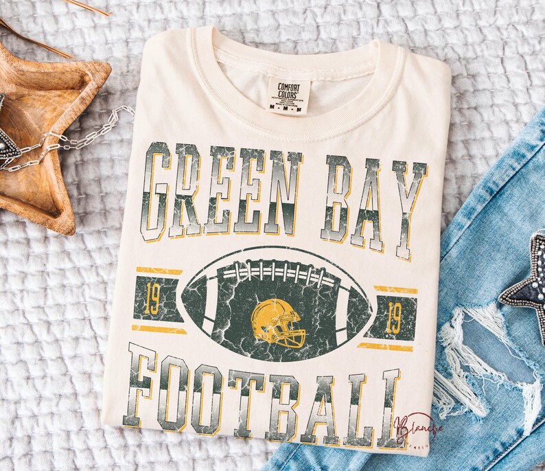 May include: Cream-coloured t-shirt featuring the text "GREEN BAY FOOTBALL" in a distressed, vintage style. A football graphic with a yellow helmet is centered below the text. The shirt has a classic crew neck and short sleeves.