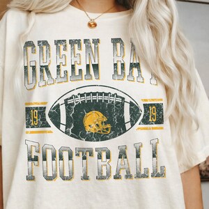 May include: Cream-coloured t-shirt with a distressed graphic design. The design features the words "GREEN BAY FOOTBALL" in a vintage font, along with a football and helmet graphic. The number "19" appears on either side of the football.
