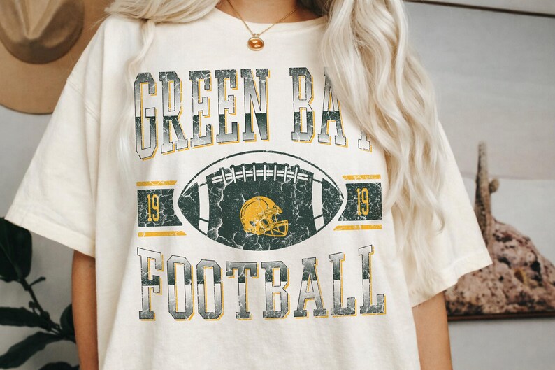 May include: Cream-coloured t-shirt with a distressed graphic design. The design features the words "GREEN BAY FOOTBALL" in a vintage font, along with a football and helmet graphic. The number "19" appears on either side of the football.