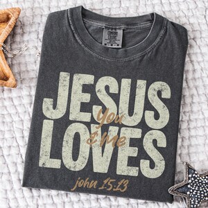 May include: Dark gray Comfort Colors t-shirt with the text "JESUS You LOVES & Me" in a distressed, off-white font. The text "john 15:13" is below. A wooden star-shaped tray and a beaded star-shaped ornament are in the background.