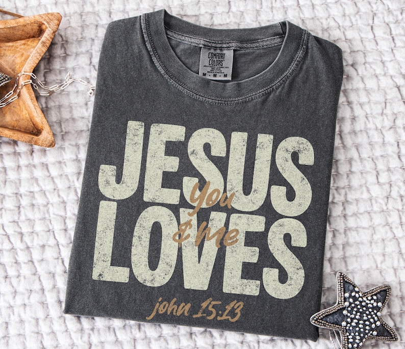 May include: Dark gray Comfort Colors t-shirt with the text "JESUS You LOVES & Me" in a distressed, off-white font. The text "john 15:13" is below. A wooden star-shaped tray and a beaded star-shaped ornament are in the background.