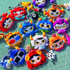 May include: A collection of colourful, cartoon-style character charms featuring various expressions, horns, and vibrant patterns. Each charm is attached to a small plastic chain, showcasing diverse designs including red, blue, orange, and purple figures.