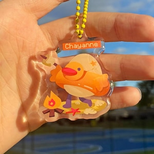 May include: A clear acrylic keychain with a cartoon illustration of a yellow bird wearing a red hat and holding a marshmallow on a stick. The bird is standing on a surfboard with a campfire and a starfish. The keychain has a yellow chain and a rectangular orange tag with the text 'Chayanne'.