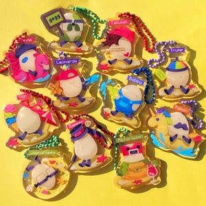 May include: A collection of 12 colorful keychains featuring cartoon characters on a yellow background. Each keychain has a different character with a name, such as 'Bobby', 'Trump', 'Tilli', 'Leonarda', 'Dapper', 'Ramon', 'Richarlison', 'Tallulah', 'Juana Flippa', '9899', 'Toque Me', and 'The character is riding a unicorn.