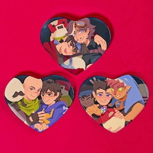 May include: Three heart-shaped pins featuring cartoon characters. The first pin shows three characters with brown hair, one wearing a red jacket and a blue hat, the second pin shows two characters with brown hair, one wearing a green shirt and a yellow scarf, and the third pin shows three characters with brown, black, and red hair, one wearing a blue shirt and a blue hat.
