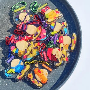 May include: A collection of colorful, cartoon-style keychains with different designs and names, including 'Juan Felipe', 'Richard', 'Dapper', 'Tilli', 'Boldboy', 'Yemile', 'Chayanne', and 'Leonarda'. The keychains are made of clear plastic with colorful designs and are arranged on a black surface.