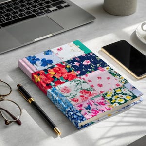 May include: A colorful patchwork journal with a floral pattern, featuring red, pink, blue, and yellow flowers. A laptop, smartphone, pen, and eyeglasses are also on the table, alongside a cup of coffee.