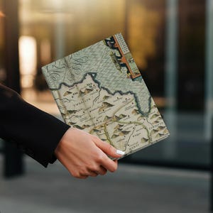 May include: A vintage-style map notebook with a detailed cartographic design. The cover features a tan and green color scheme with intricate illustrations of mountains, rivers, and text. The notebook is held in a person's hand.