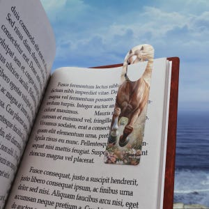 Può includere: Un segnalibro con un'illustrazione di un cavallo in corsa. Il segnalibro è inserito in un libro aperto, con testo visibile sulle pagine. Il cavallo è marrone e bianco, con un motivo floreale nella parte inferiore.