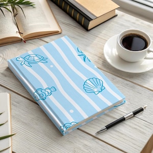 Könnte beinhalten: Ein blau-weiß gestreiftes Notizbuch mit Illustrationen von Meeresbewohnern, darunter eine Schildkröte, ein Fisch und eine Muschel, liegt auf einem Holztisch. Eine Tasse Kaffee, ein Stift und offene Bücher befinden sich ebenfalls auf dem Tisch.